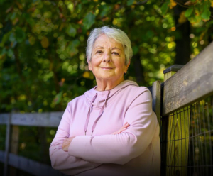 Smiling elderly woman leaning against a fence under the trees.