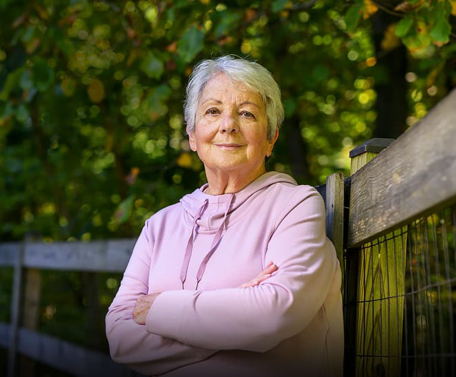 Smiling elderly woman leaning against a fence under the trees.